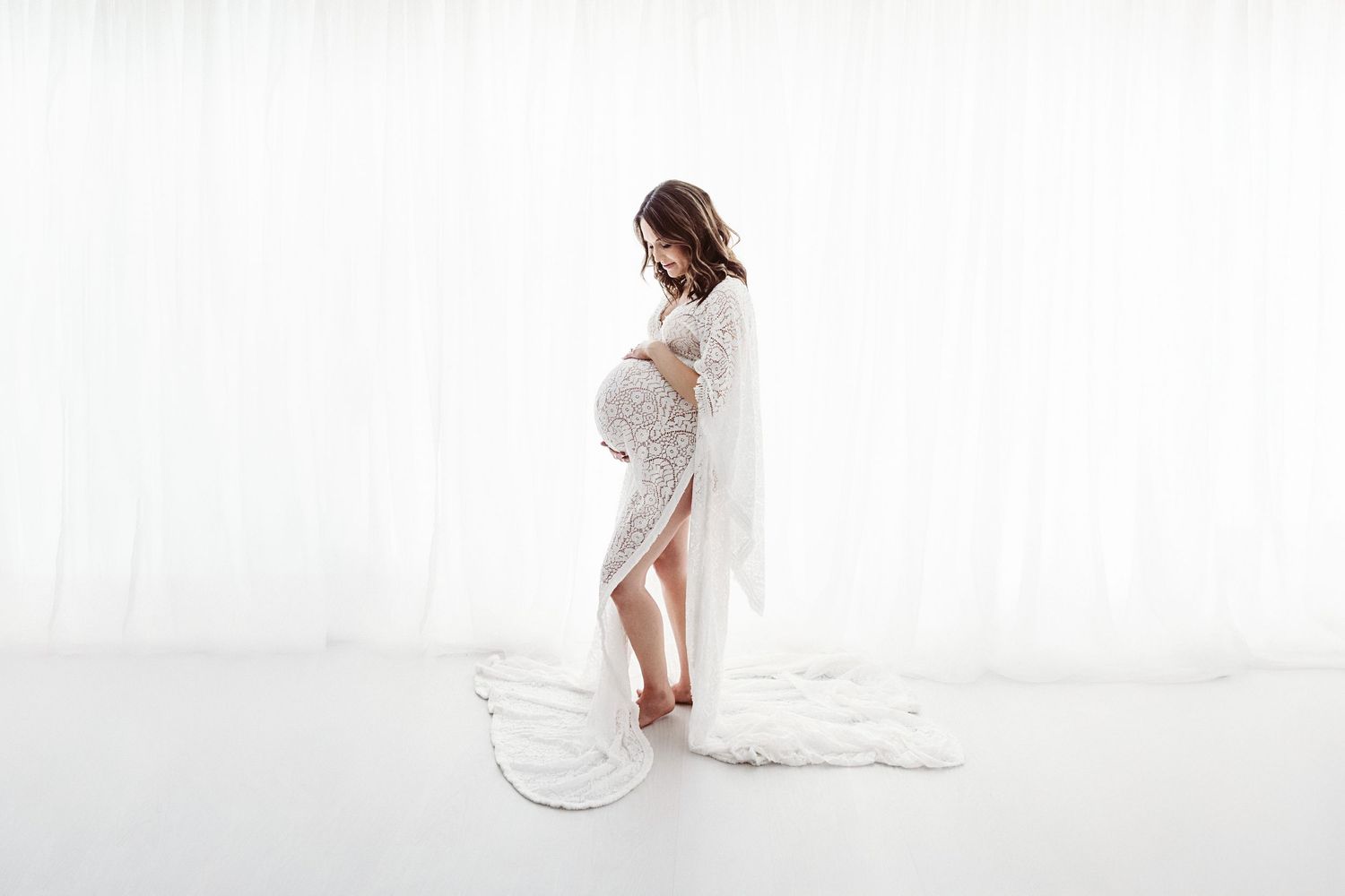 Pregnant woman in a white lace maternity gown posing gracefully against a white backdrop.
