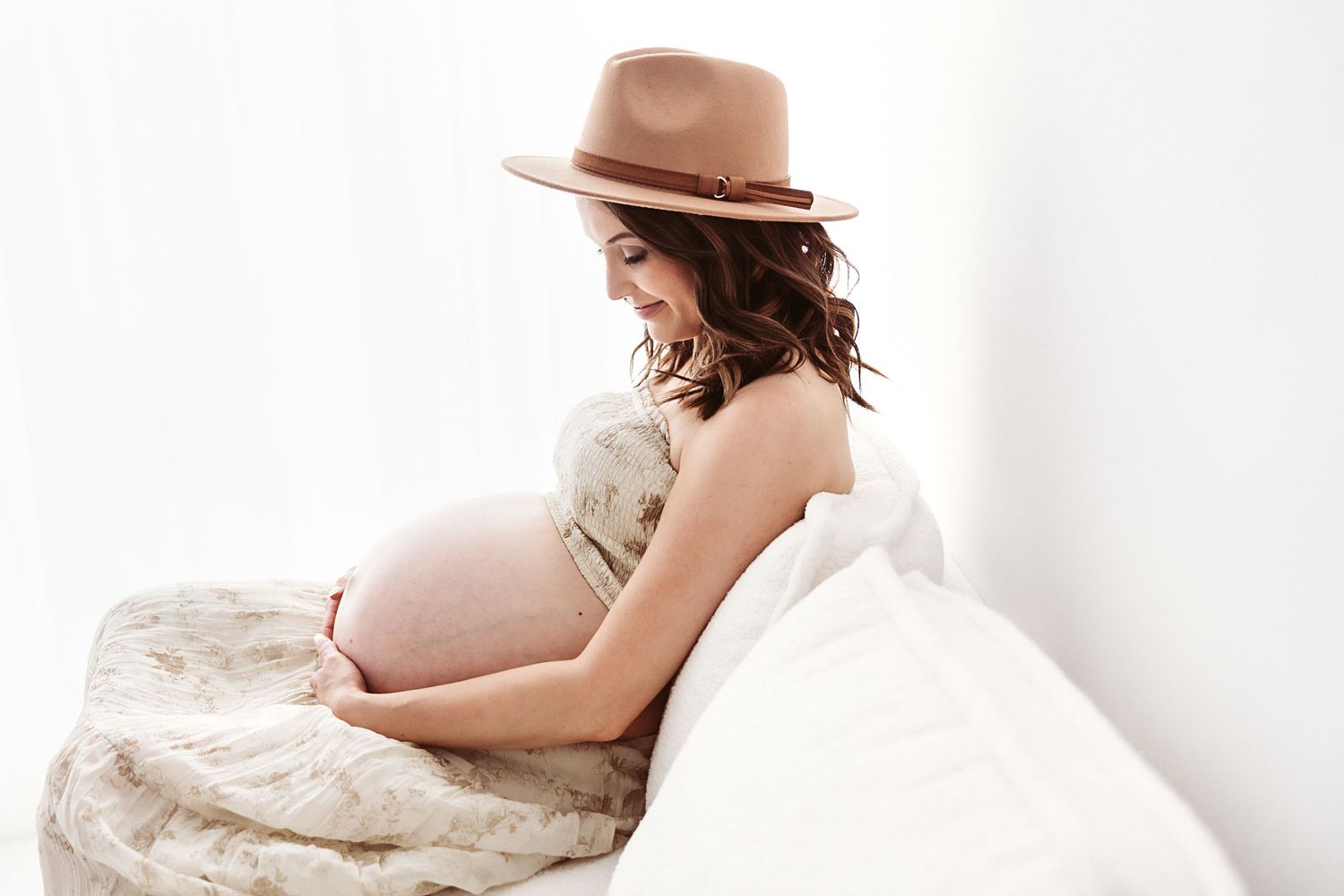 Pregnant woman in boho hat and floral dress sitting on white bed holding baby bump in studio.