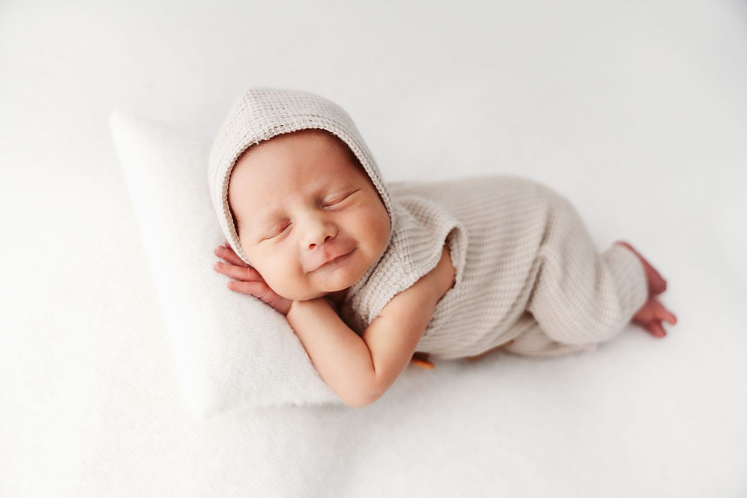 Adorable sleeping newborn wearing beige hooded knit outfit resting on white pillow.