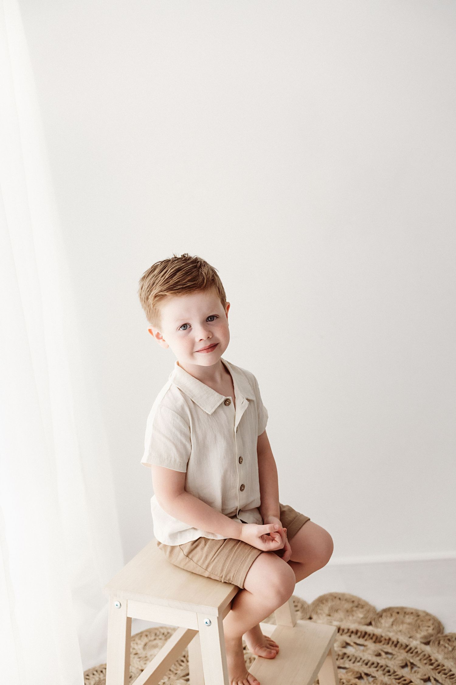 A cute young boy in a beige linen shirt and khaki shorts sits on a wooden stool in a bright studio.
