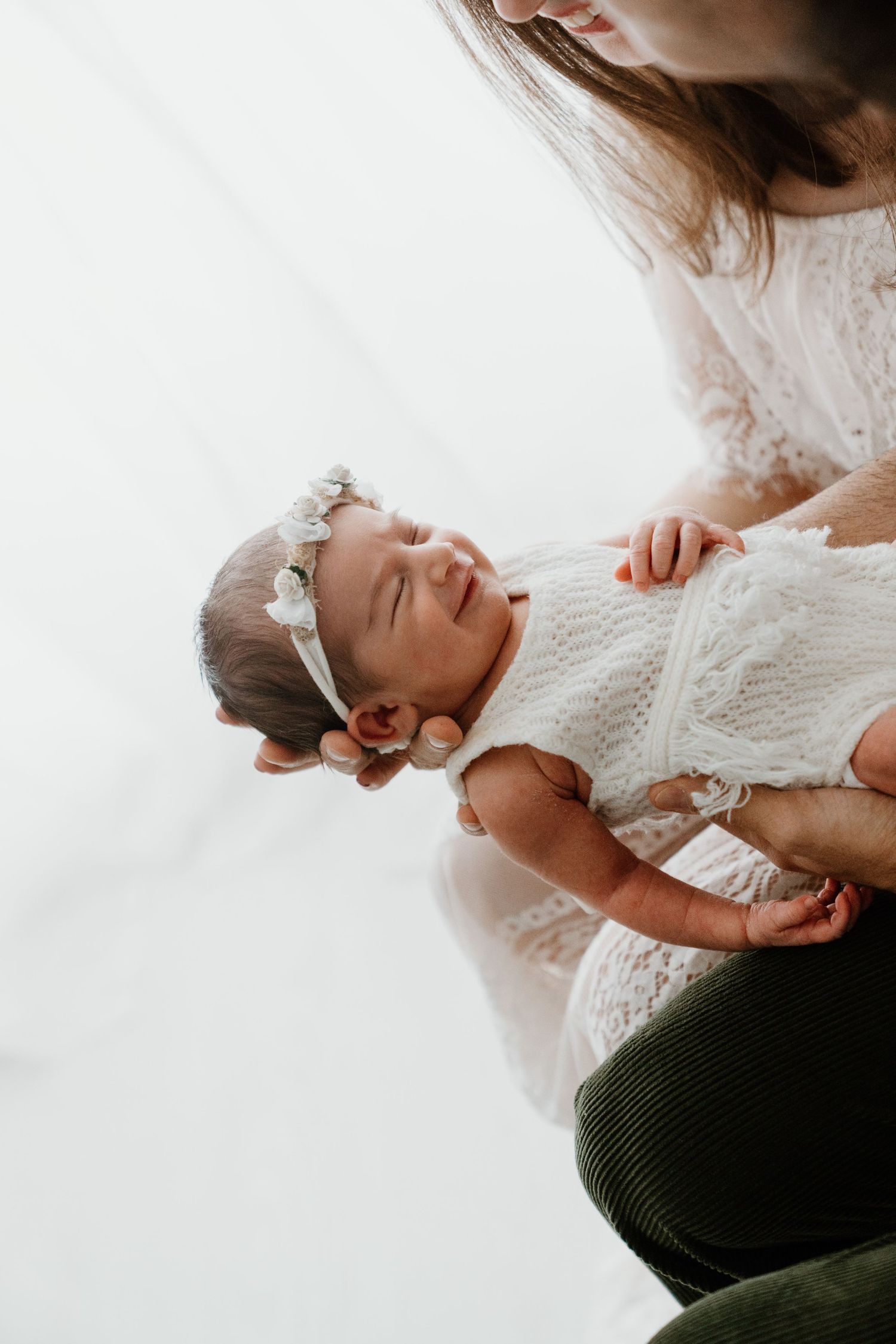 Mother in white lace dress gently holding sleeping newborn with floral headband.