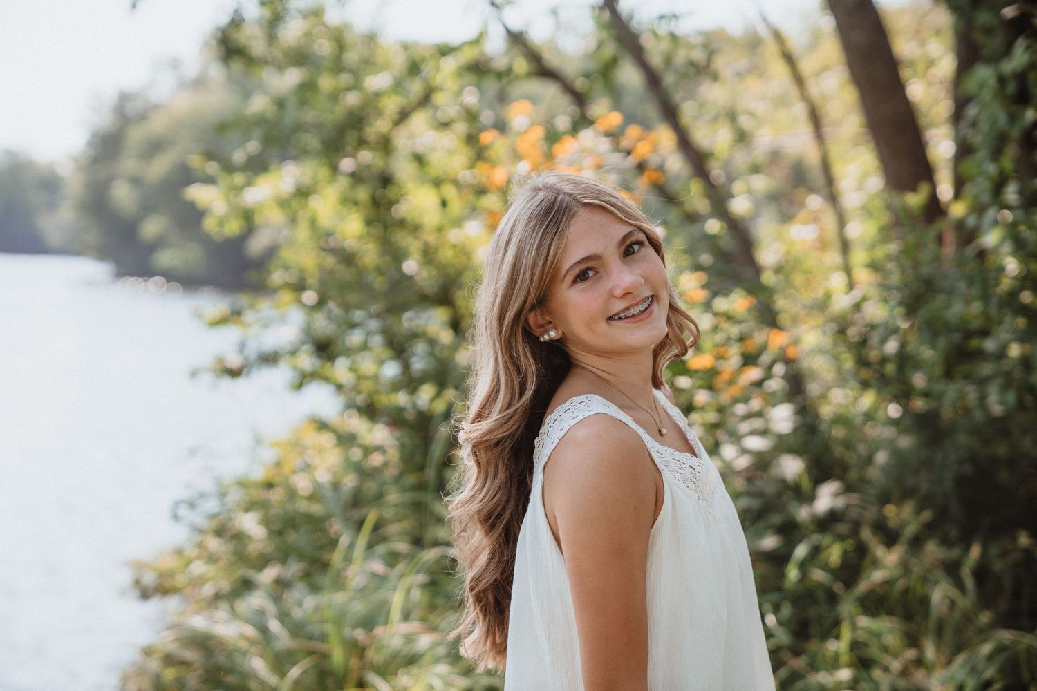 Young woman in white dress smiling outdoors near a lake surrounded by lush green trees in warm sunlight.