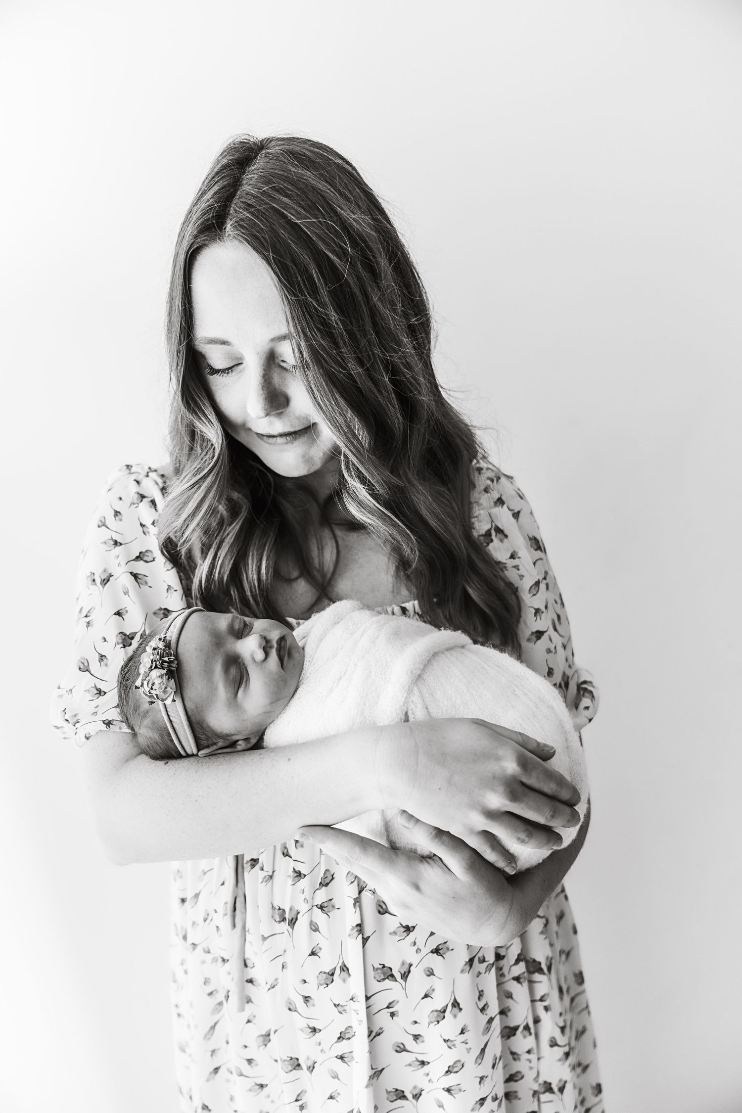 A black and white studio portrait of a mother in a floral dress lovingly holding her swaddled newborn.
