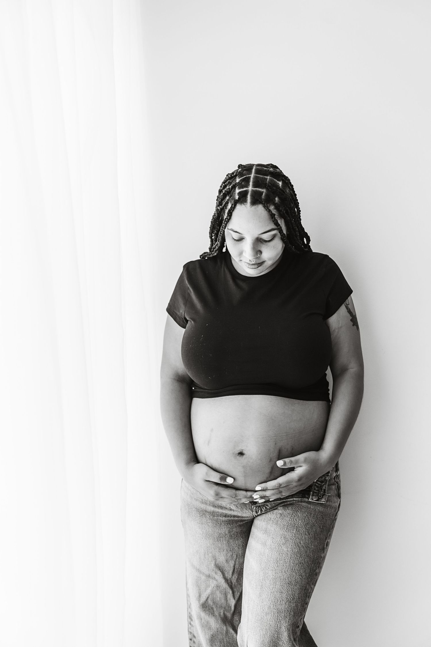 Black and white photo of pregnant woman in black top holding her bare baby bump near a curtain.