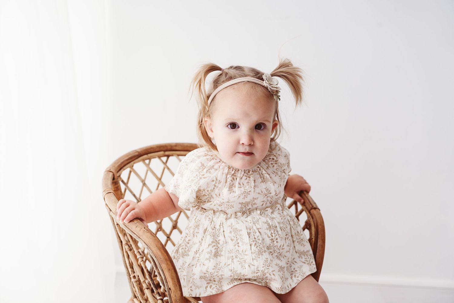 Adorable baby girl with pigtails in floral romper sitting in a wicker rattan chair.