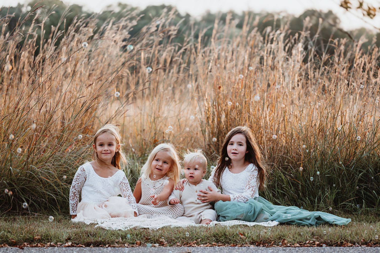 Four young children sitting together on blanket near tall golden grass field.