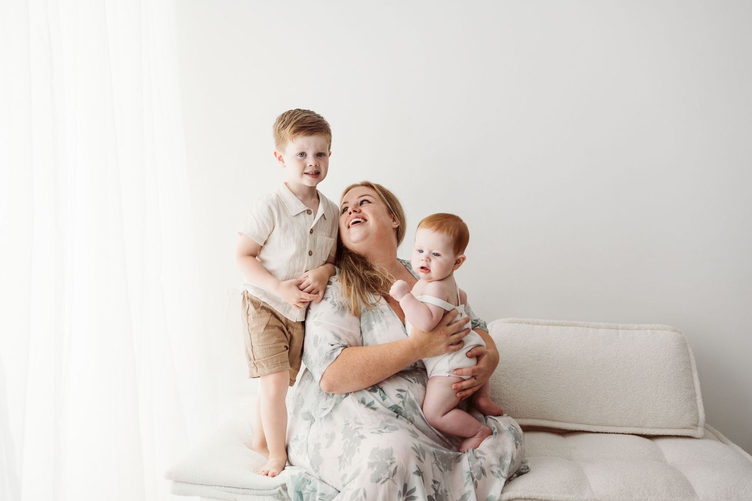 A joyful mother sits on a white sofa laughing with her toddler son and baby in a bright studio.