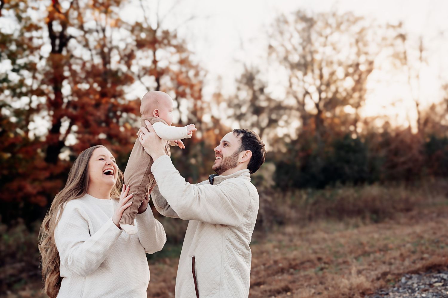 Laughing parents lifting their baby in the air outdoors during a fall family photo session.