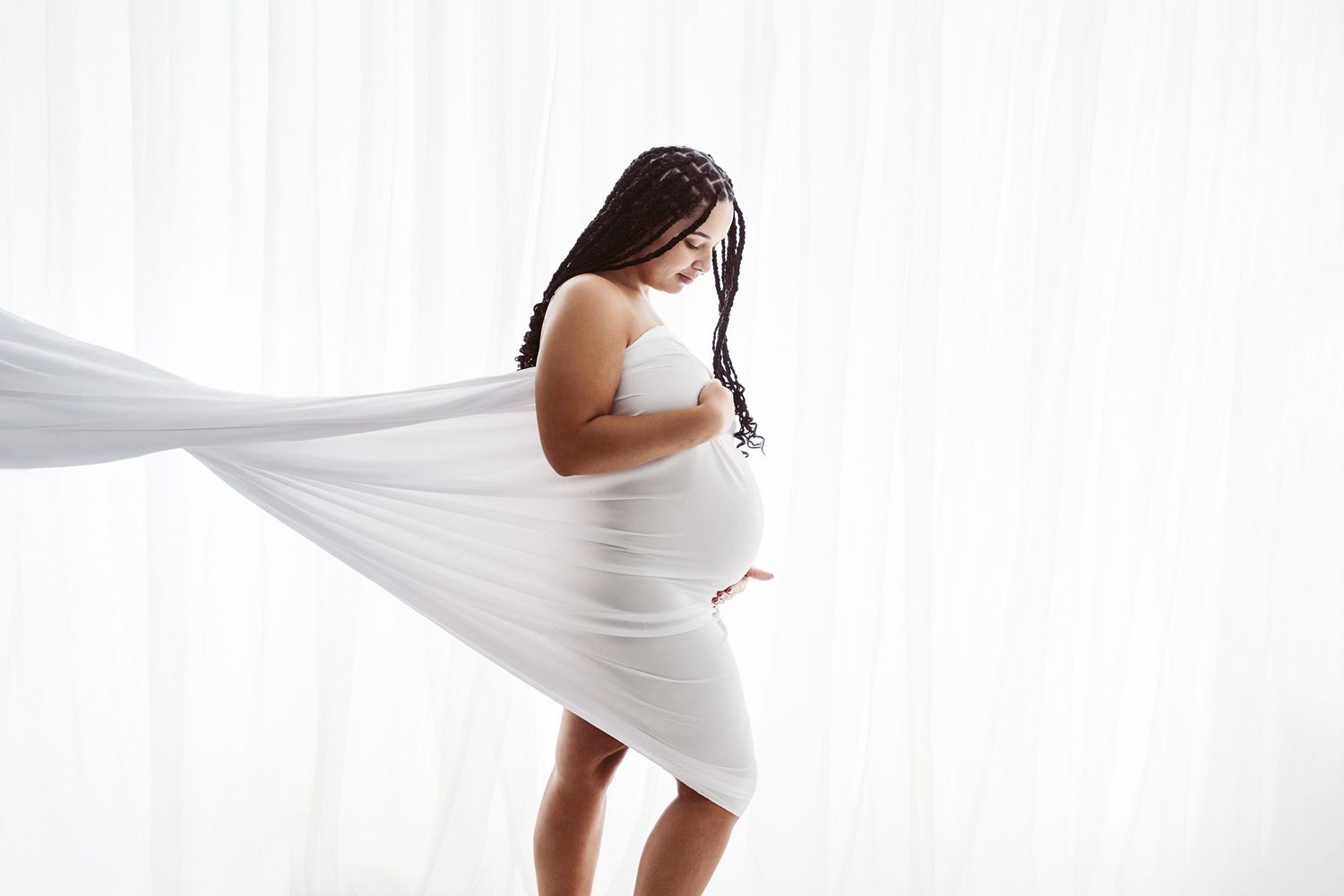 Pregnant woman with braids holding white flowing fabric around her bump in a bright white studio.