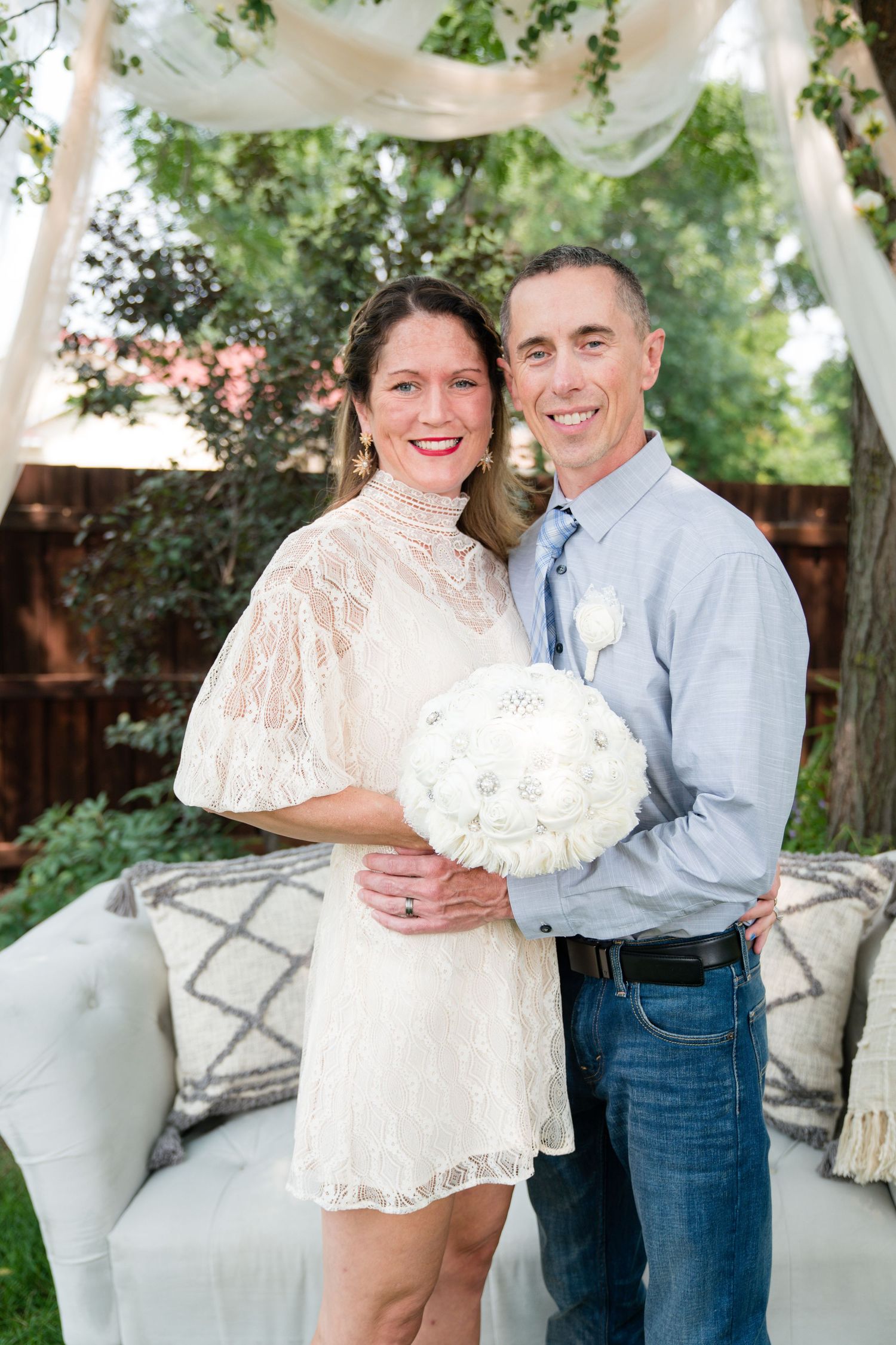 Happy wedding couple posing outdoors with white bouquet, lace dress, and blue tie in garden setting.