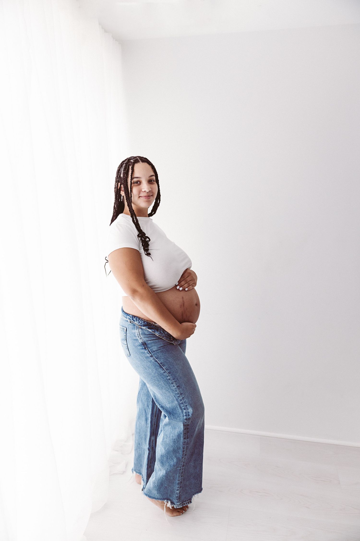 Smiling pregnant woman in white top and blue jeans standing sideways cradling her bump by curtains.