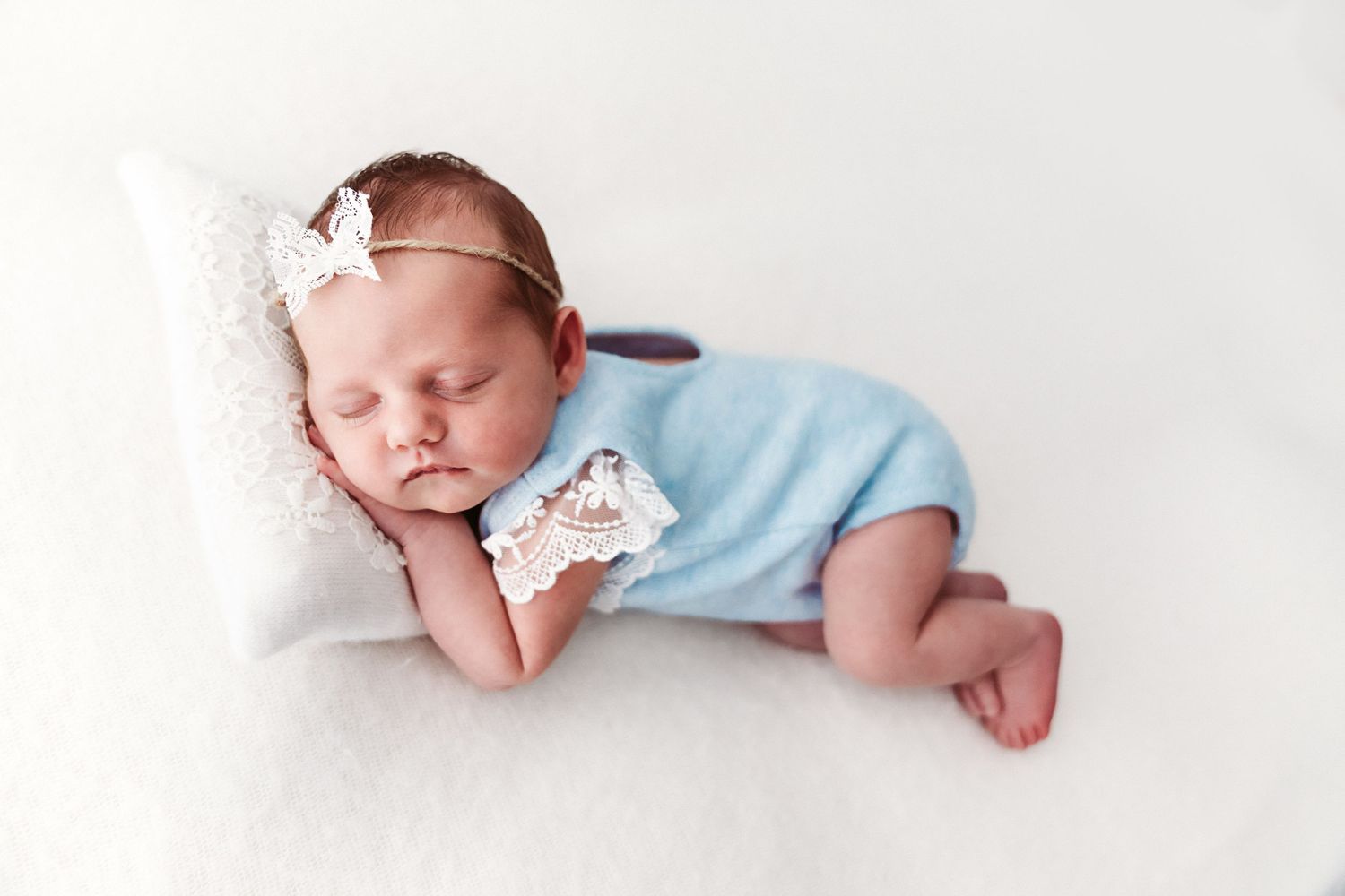 Newborn baby girl sleeping in blue romper with lace bow headband on white blanket.