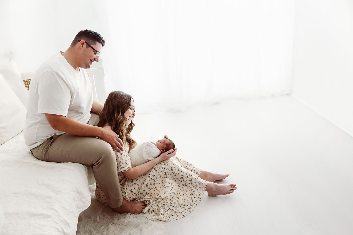 A family sits on a white bed, parents lovingly gazing at their newborn baby in a bright, airy room.