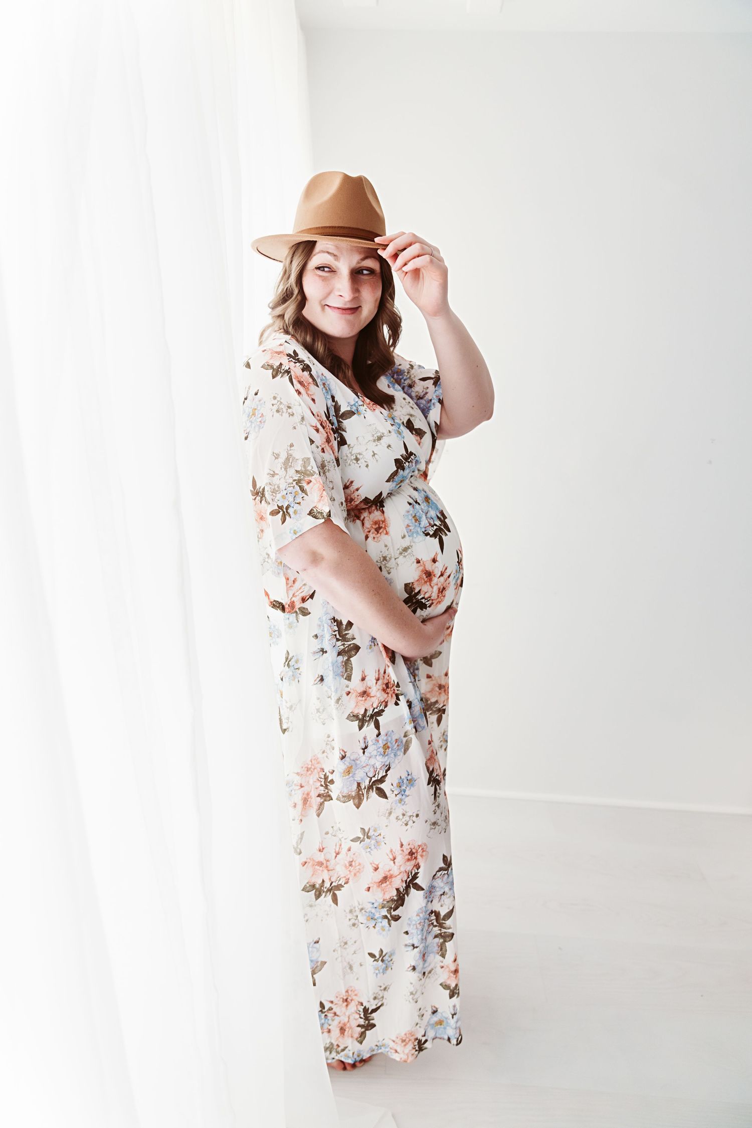 Smiling pregnant woman in floral dress and brown hat posing in a bright white photography studio.