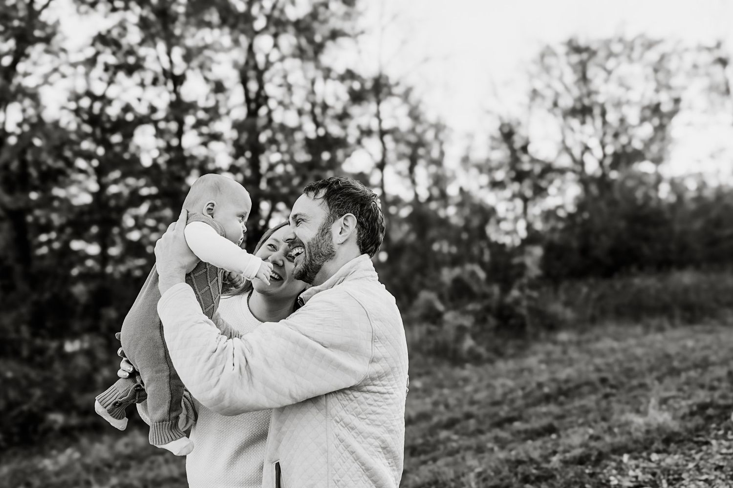 Black and white photo of a smiling father lifting his laughing baby outdoors among trees.