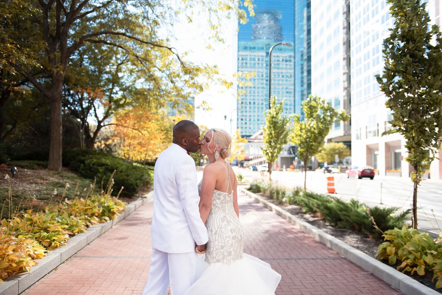 Interracial newlyweds walking hand in hand on urban sidewalk, groom in white suit, bride in beaded gown.