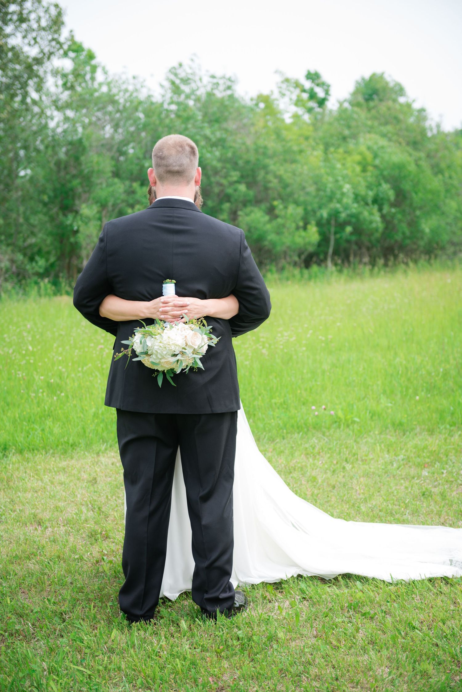 Groom holding a white bridal bouquet behind his back while standing in a lush green field.