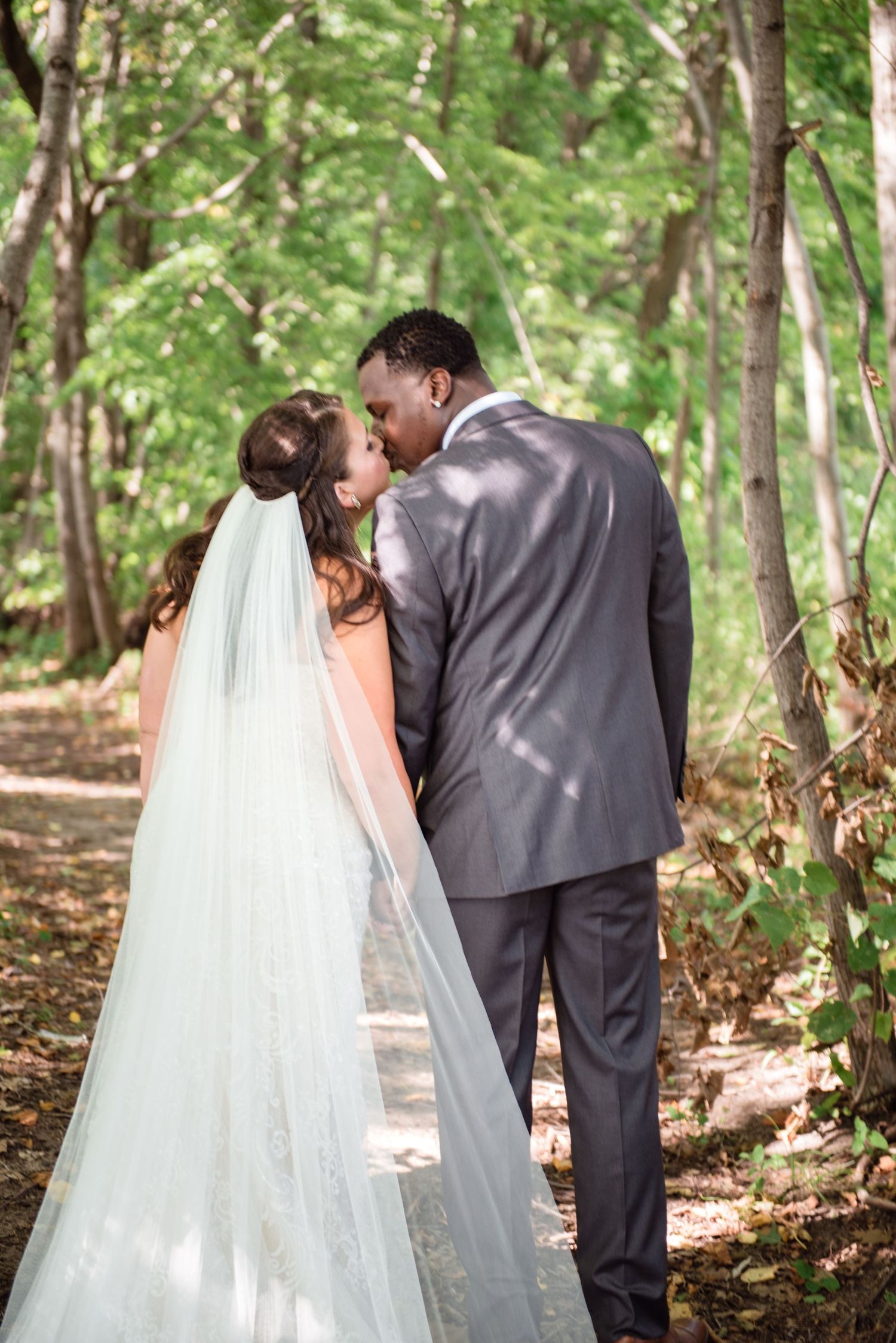 Bride and groom share a kiss on a sunlit wooded path, veil trailing behind.