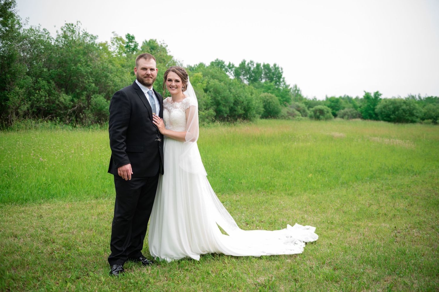 Bride and groom posing together in a green meadow, bride wearing a flowing white gown with train.
