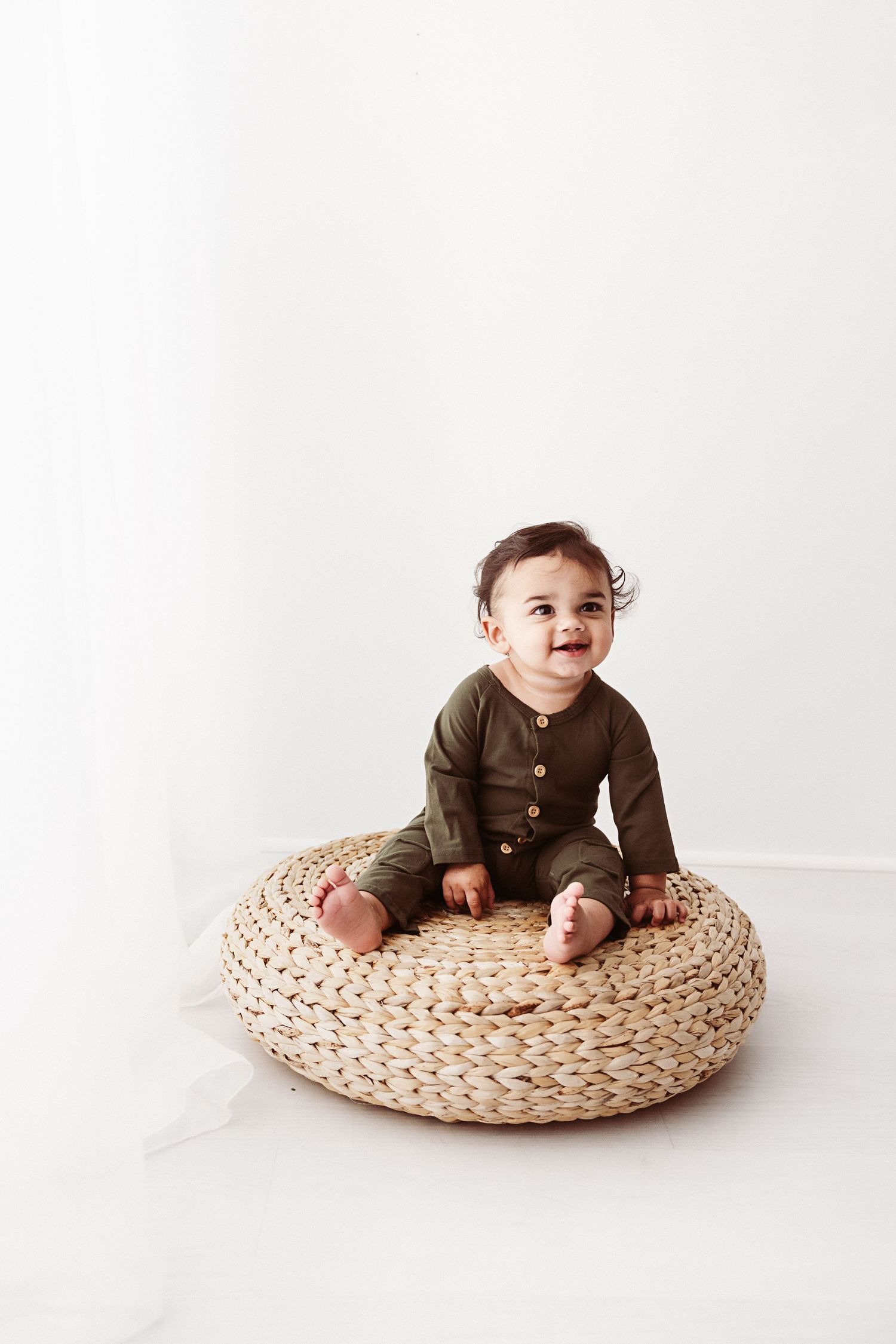 Happy baby in olive green romper sitting on a woven rattan pouf against a white background.