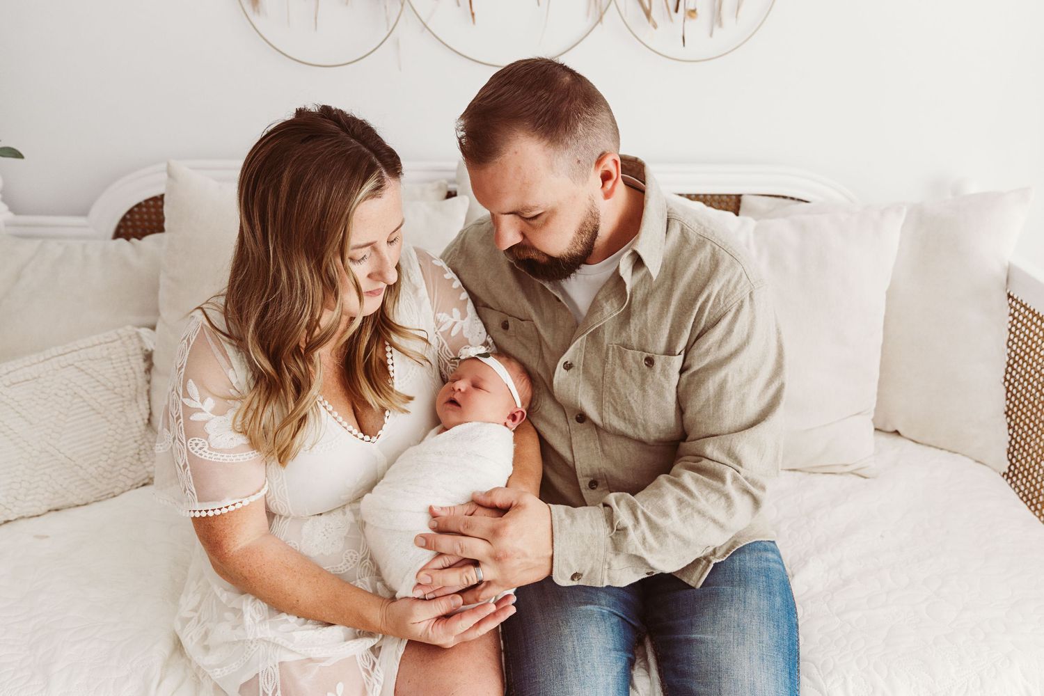 Happy parents sitting on white bed smiling at their swaddled newborn baby in a bright studio setting.