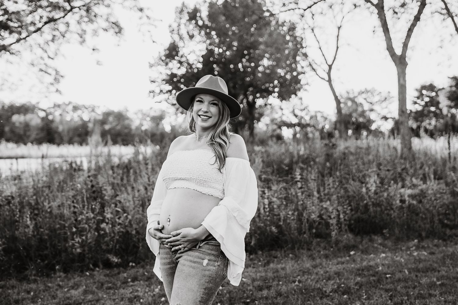 Black and white maternity photo of a smiling pregnant woman in a hat and jeans in an outdoor field setting.