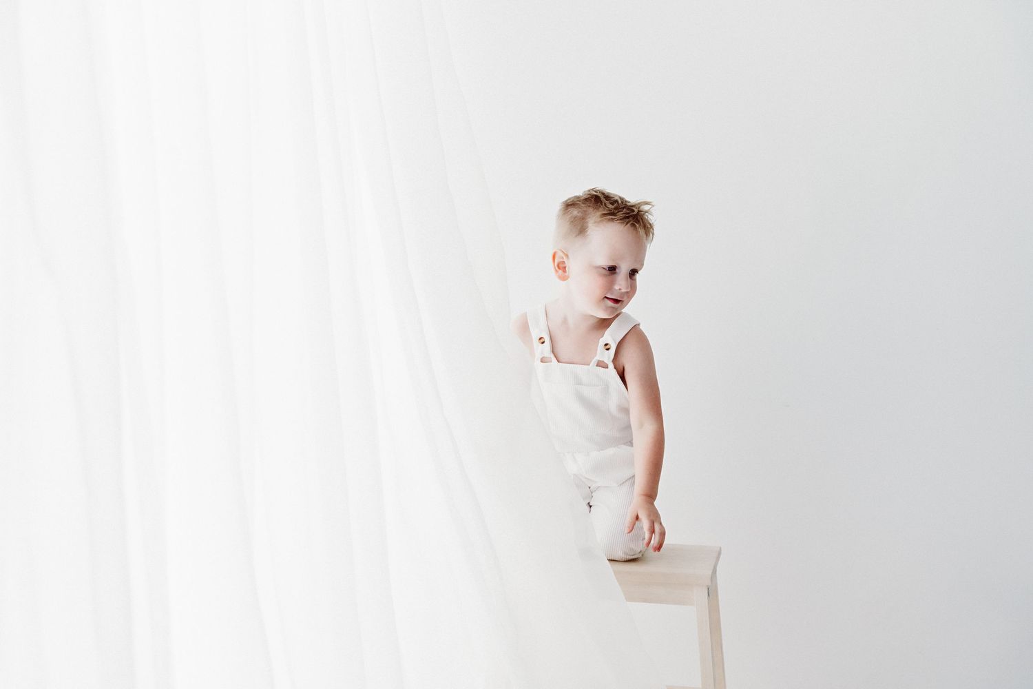 Toddler boy in white overalls standing behind a wooden chair against a white curtain backdrop.