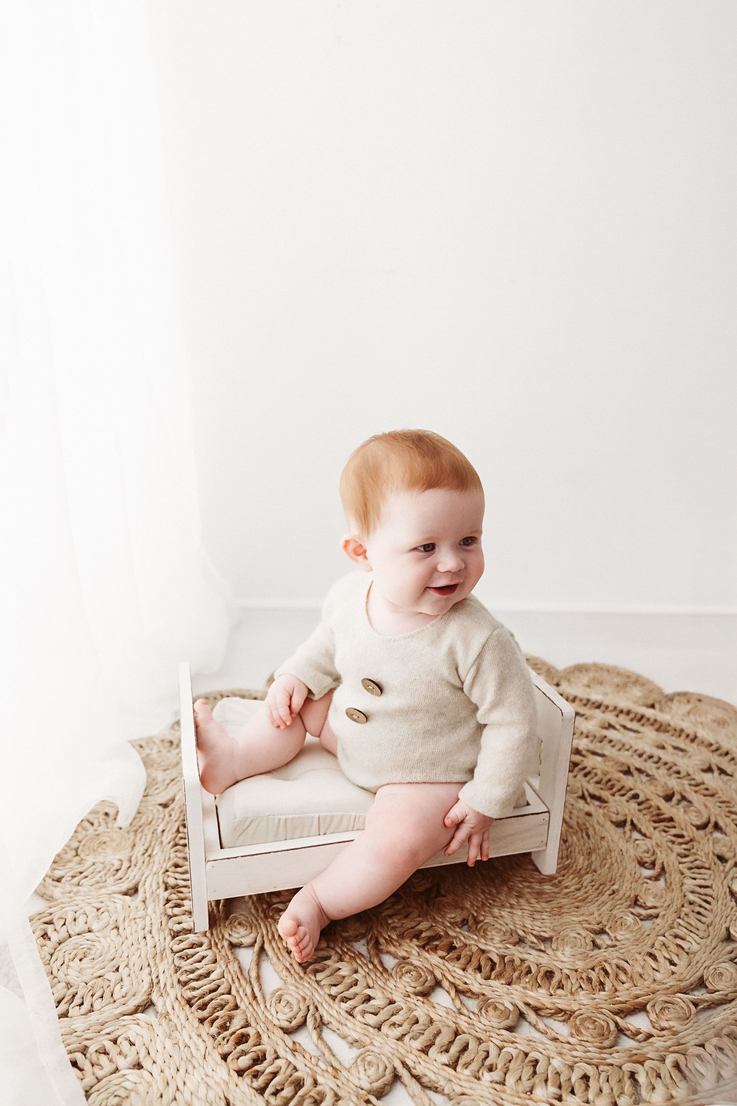 An adorable red-haired baby in a cream knit romper sits on a miniature white bed on a jute rug.