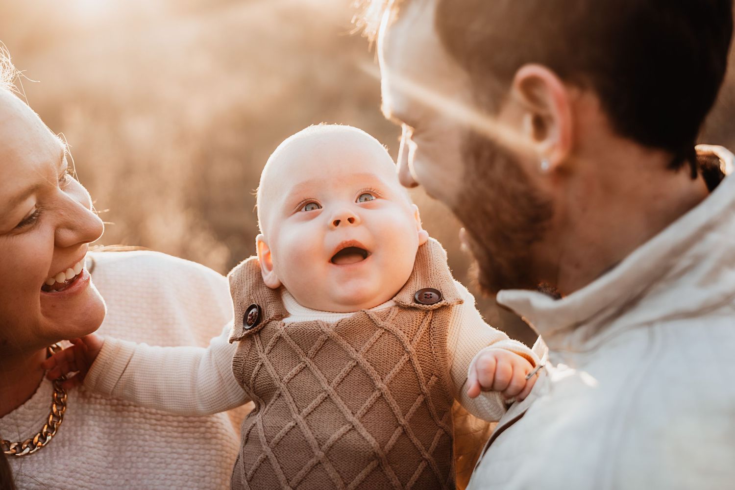 Adoring parents smiling at their bright-eyed baby in warm golden sunset light outdoors.