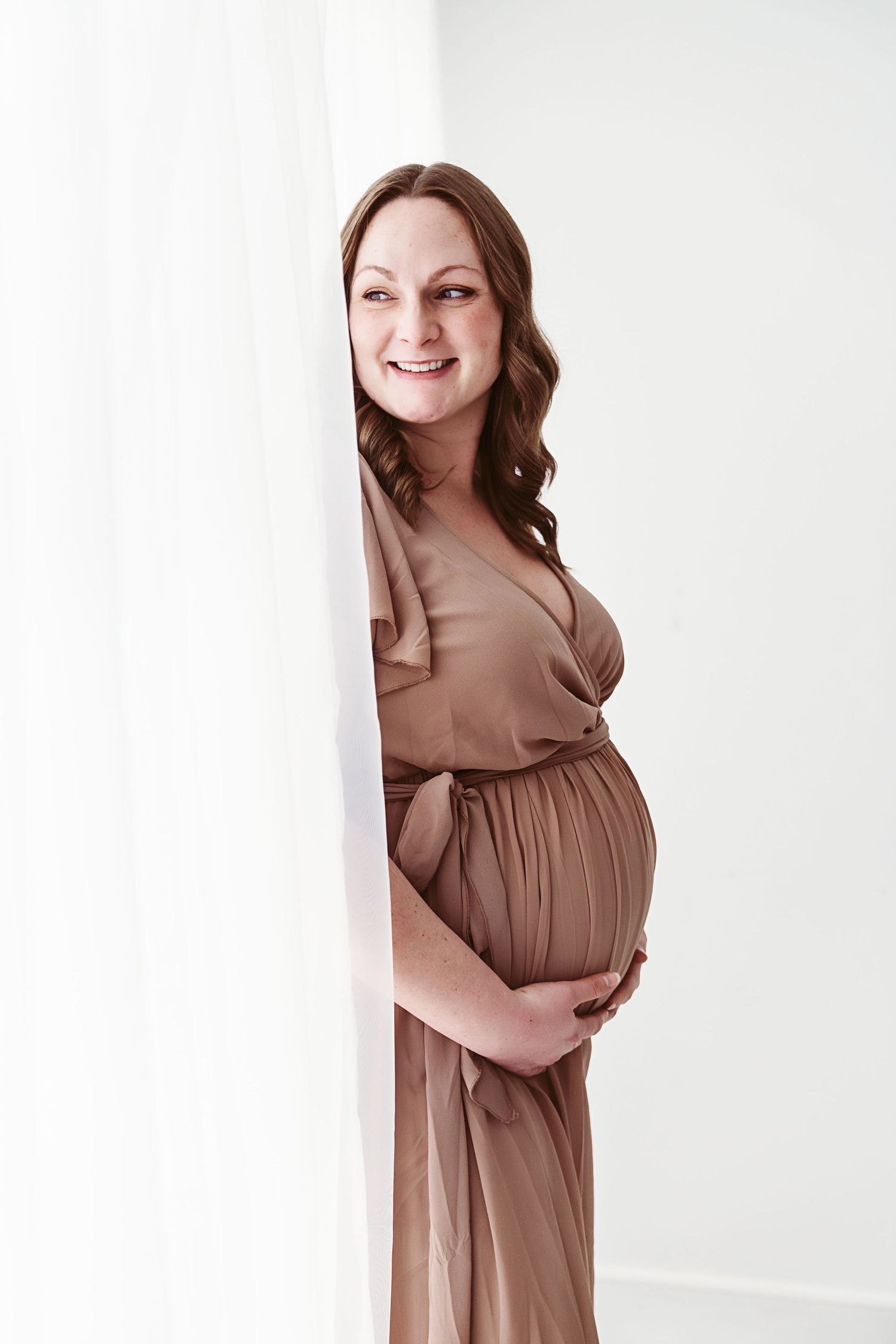 Smiling pregnant woman in tan wrap dress peeking around white curtain holding baby bump.