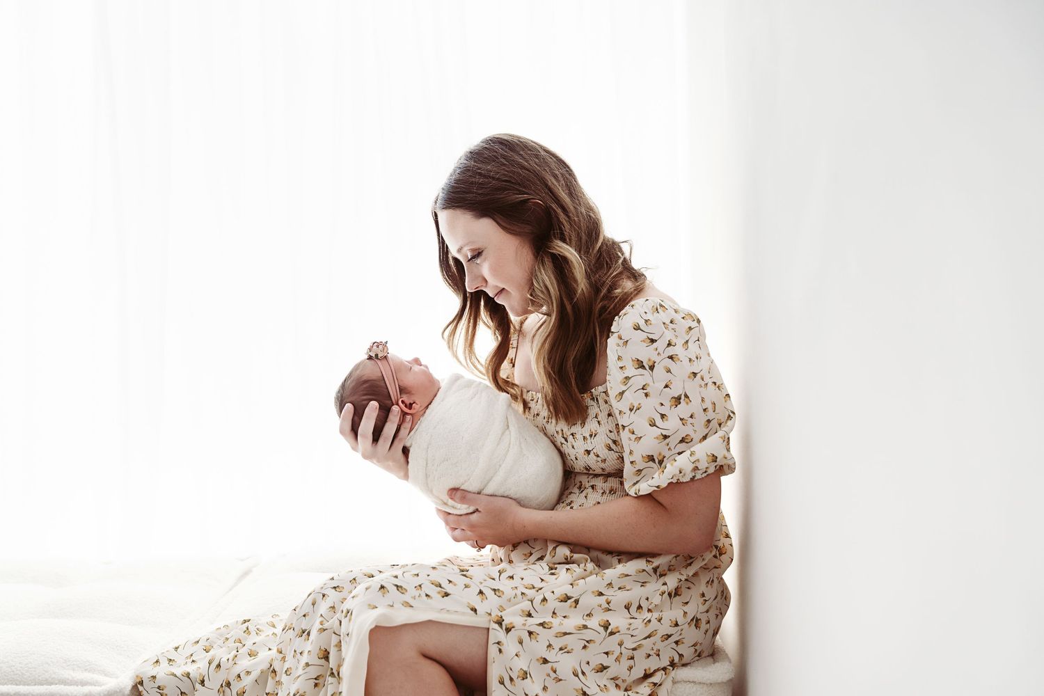 A brunette mother in a floral dress sits by a window, lovingly cradling her swaddled newborn.