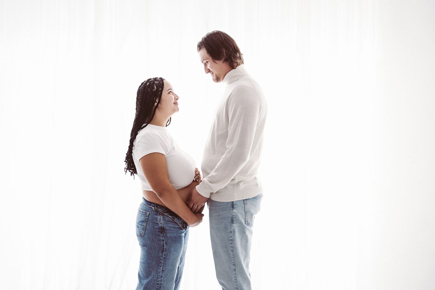 Expecting couple facing each other holding pregnant belly together against white curtain backdrop.