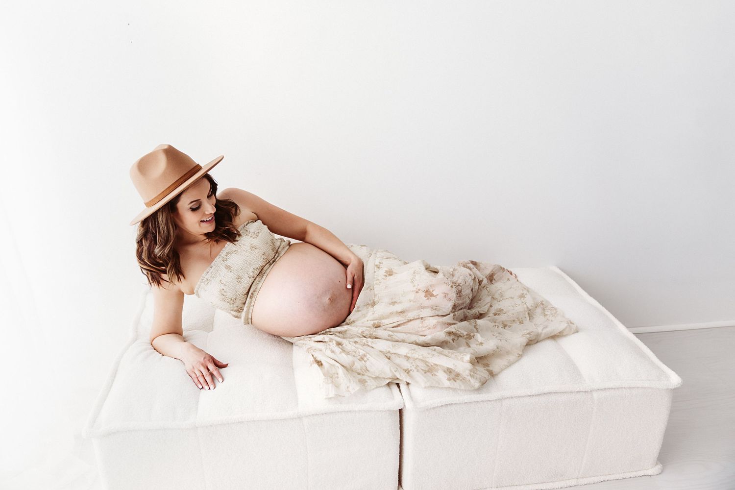 Pregnant woman in a floral dress and tan hat reclining on a white bed.