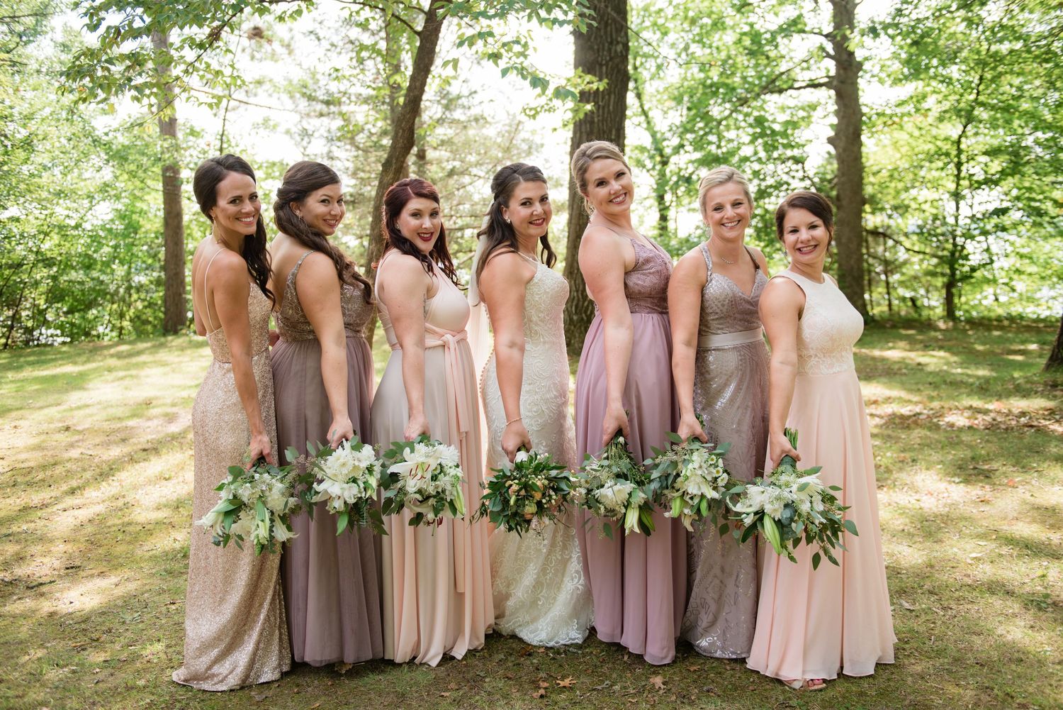 Seven bridesmaids in mismatched blush and taupe gowns pose with white bouquets in a wooded outdoor setting.