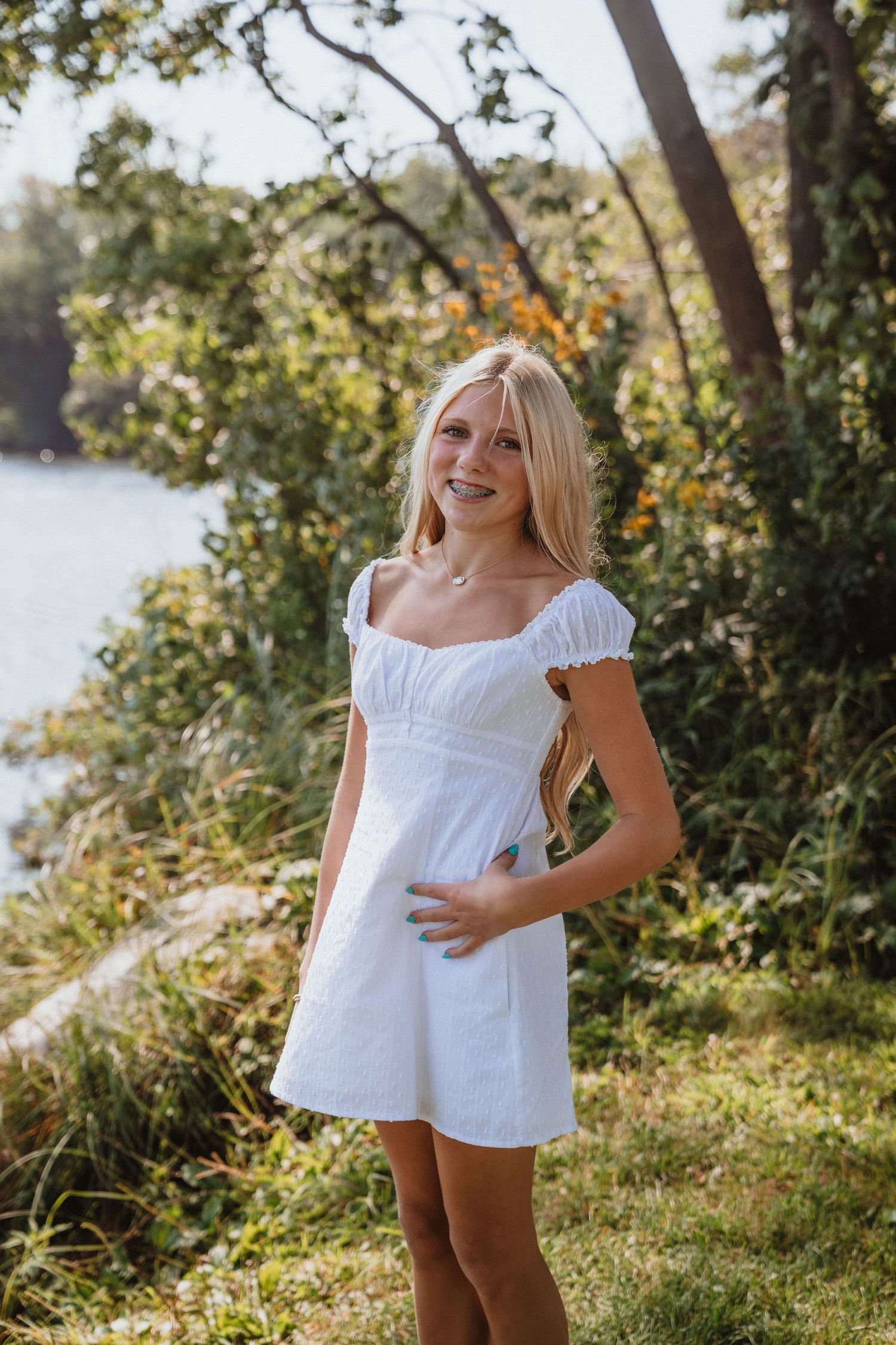 Young blonde woman in white mini dress smiling outdoors near a lake surrounded by lush green trees.