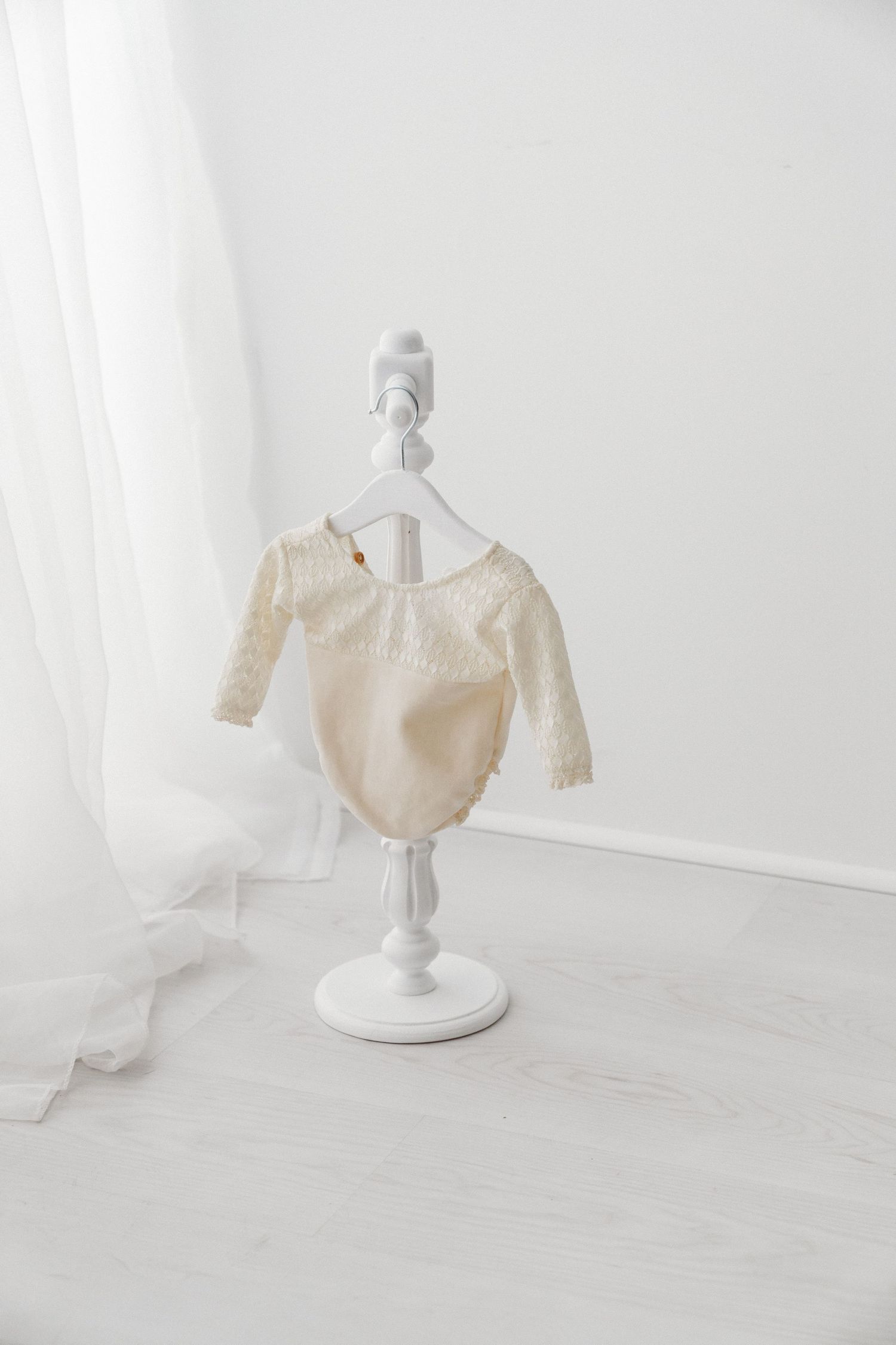 Elegant cream lace baby outfit with bloomers hanging on a white decorative mini stand in a bright white studio room.