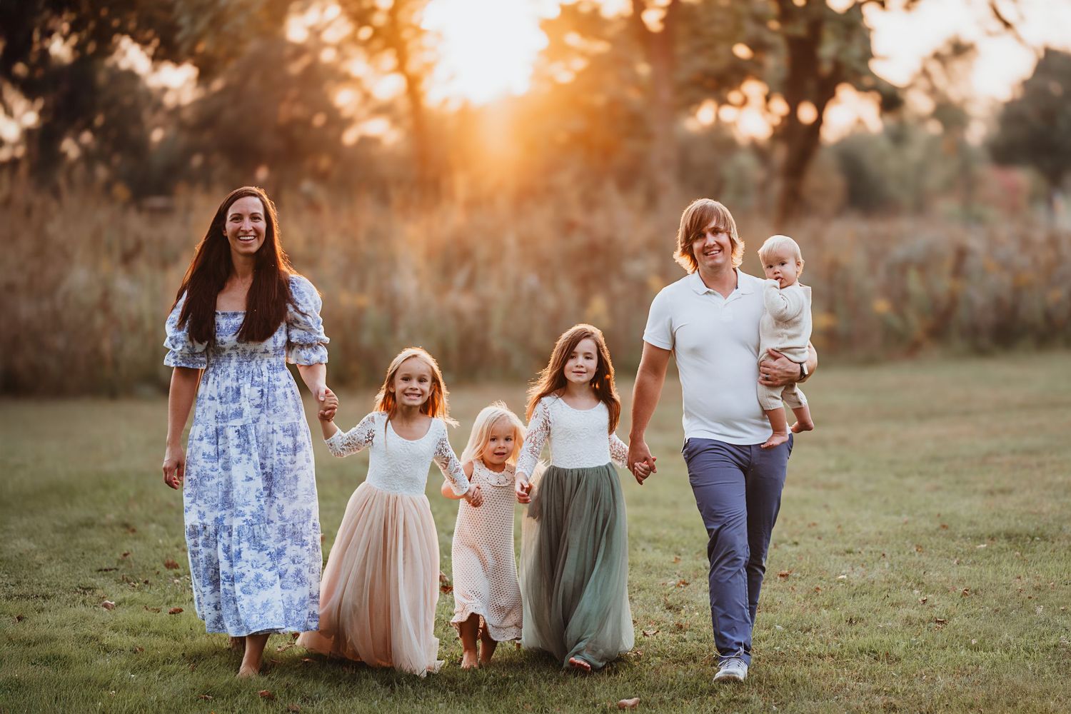 A happy family of six walks together through a sunlit green meadow at sunset.