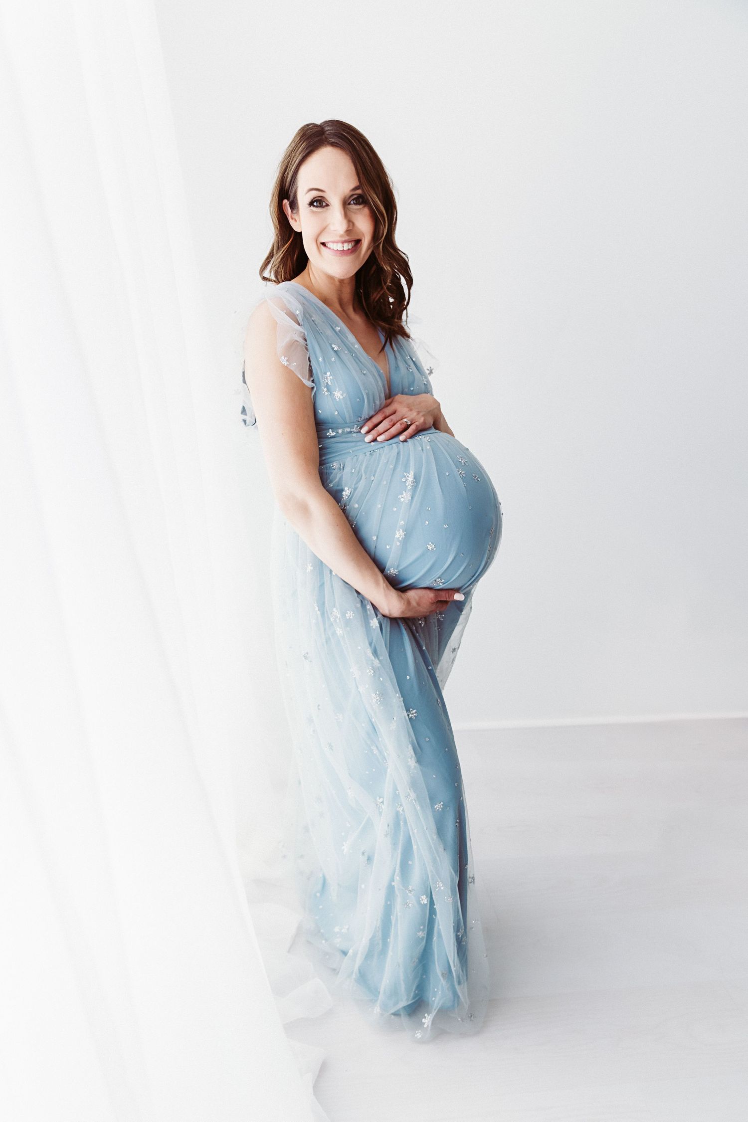 A smiling pregnant woman in a blue star-embroidered tulle gown cradles her baby bump in a white studio.
