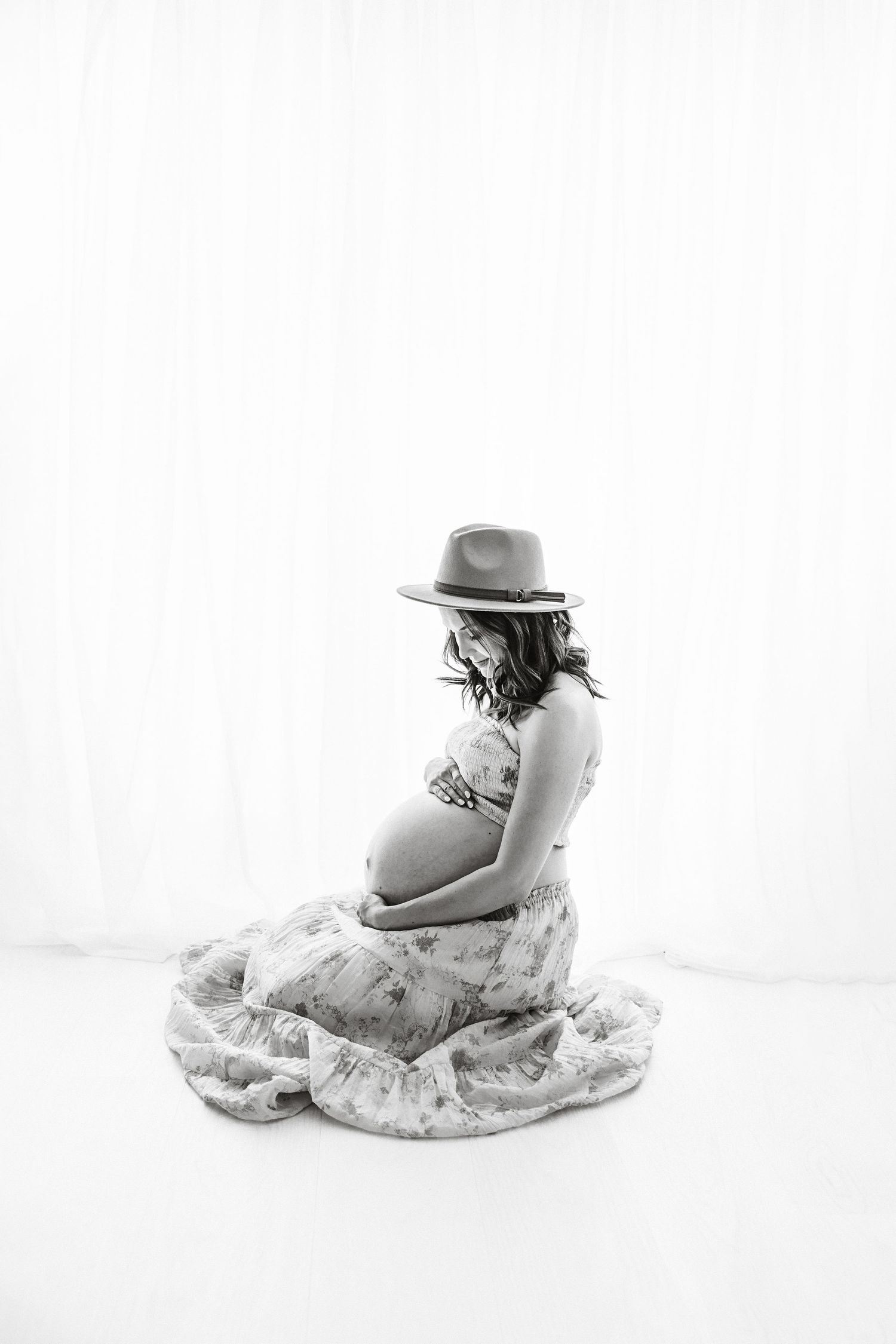 Pregnant woman in floral dress and hat sitting gracefully on floor, black and white maternity photo.