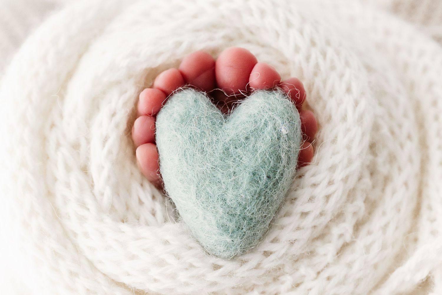Newborn baby feet holding a soft mint green felted wool heart wrapped in a white knit blanket.