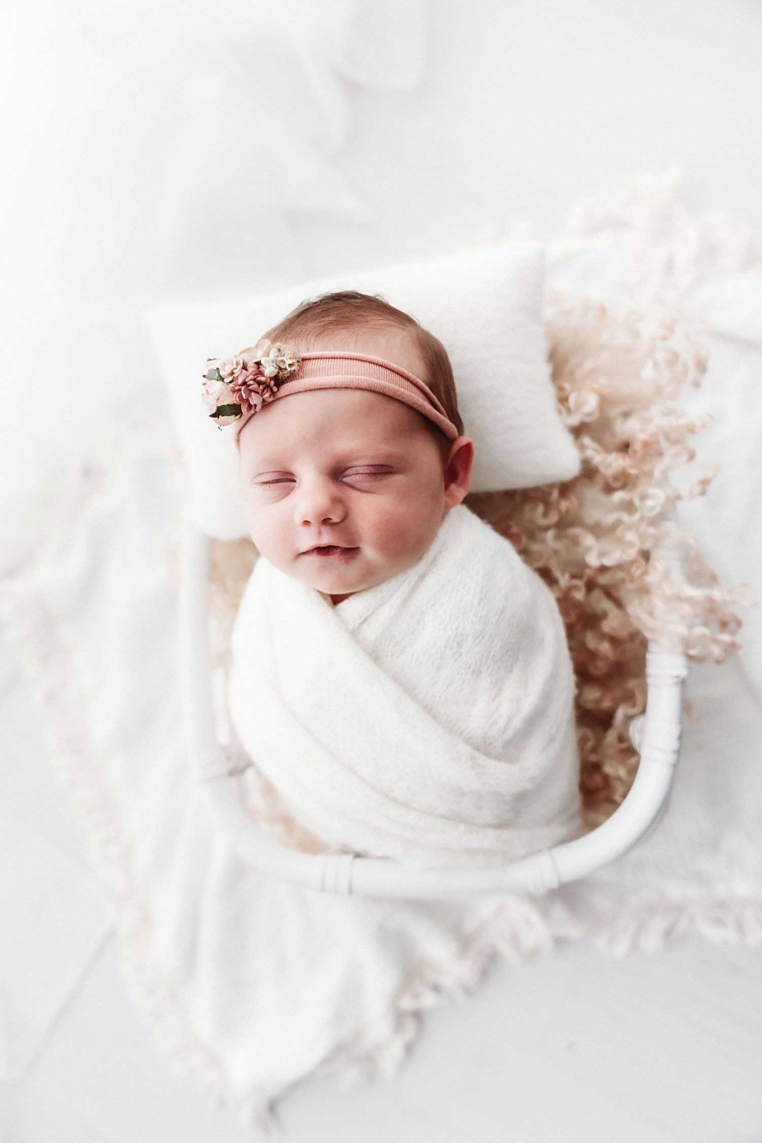 A swaddled newborn baby wearing a pink floral headband sleeps in a tiny white bed with soft blankets.