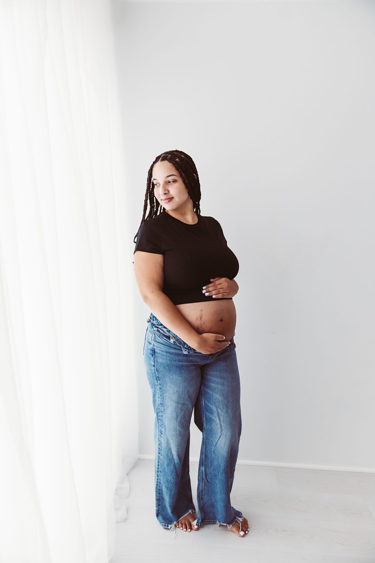 Pregnant woman in black top and jeans cradling her baby bump while standing near a white curtain.