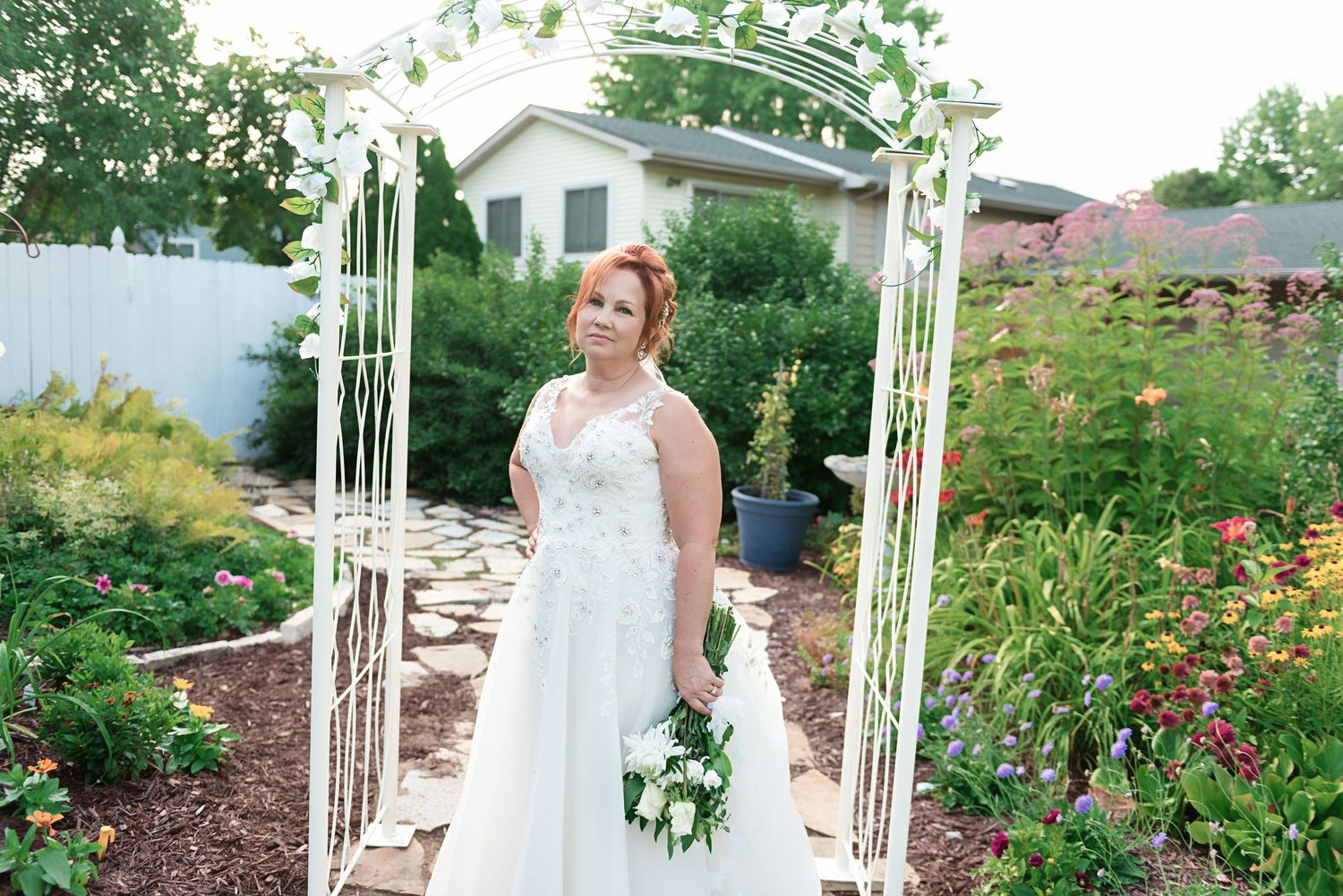 A red-haired bride in a white lace gown stands under a floral arch in a colorful garden.