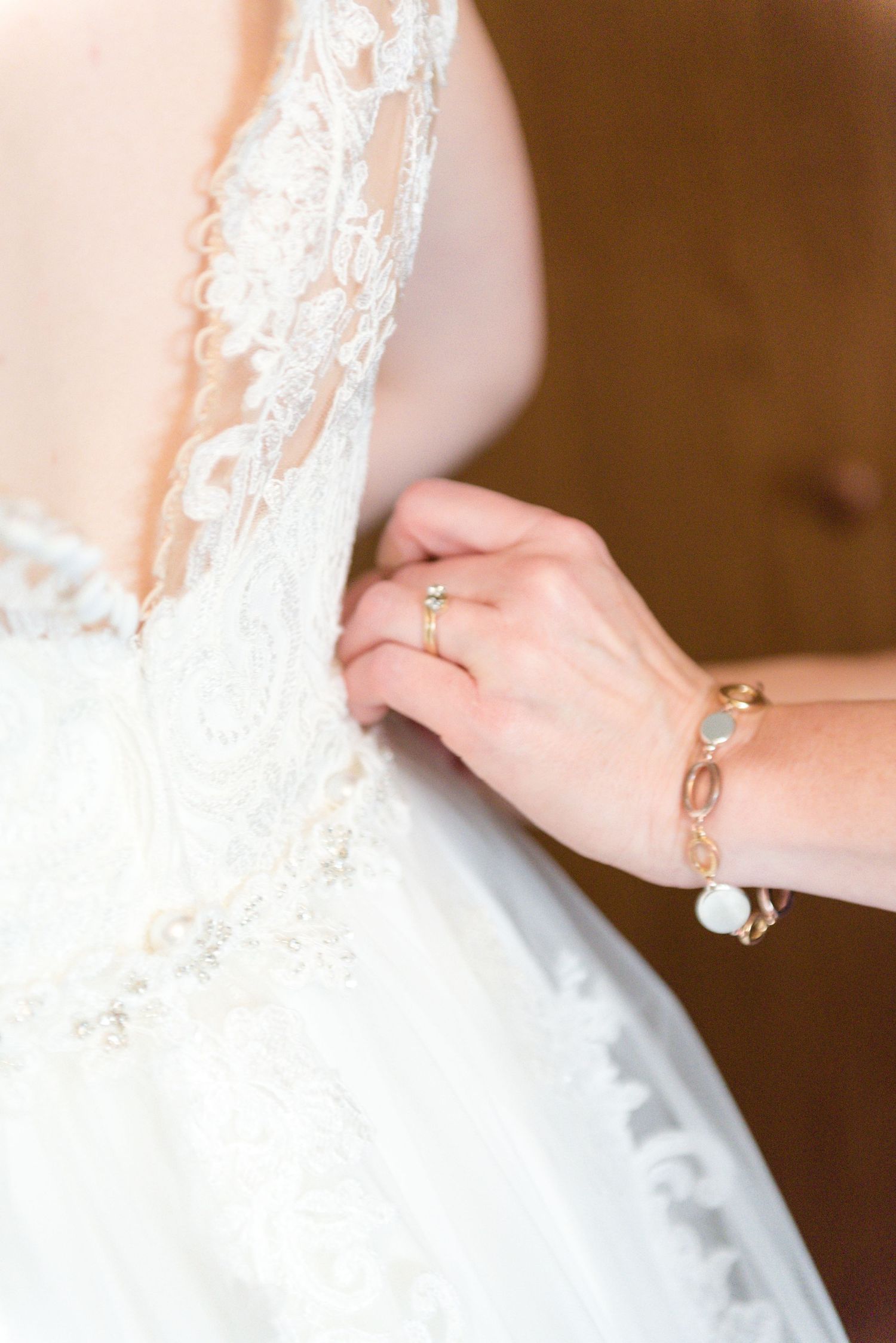 Close-up of hands fastening a lace wedding dress, adorned with a gold ring and bracelet.