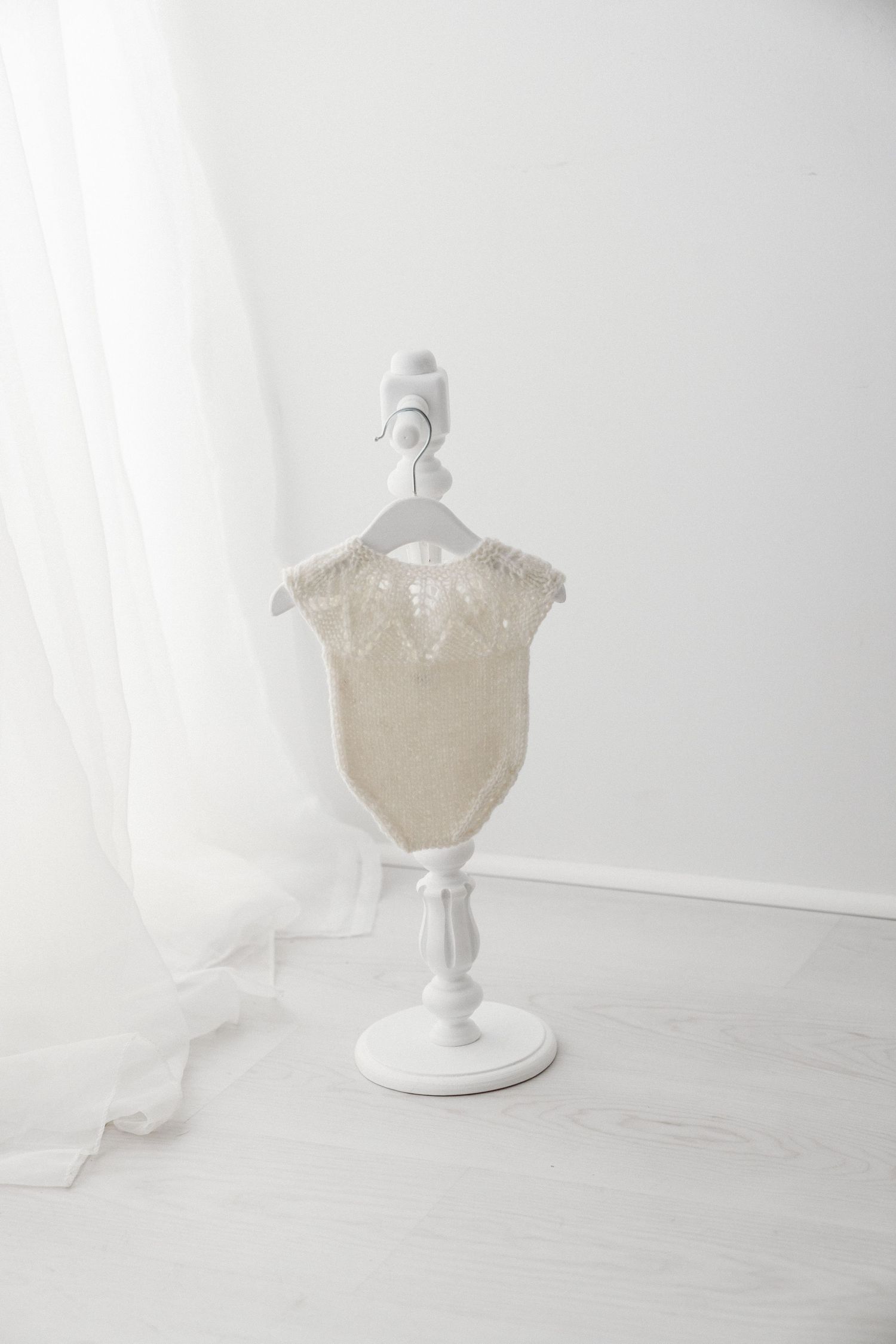 Cream knitted baby romper with delicate lace detail hanging on a white decorative mini stand.