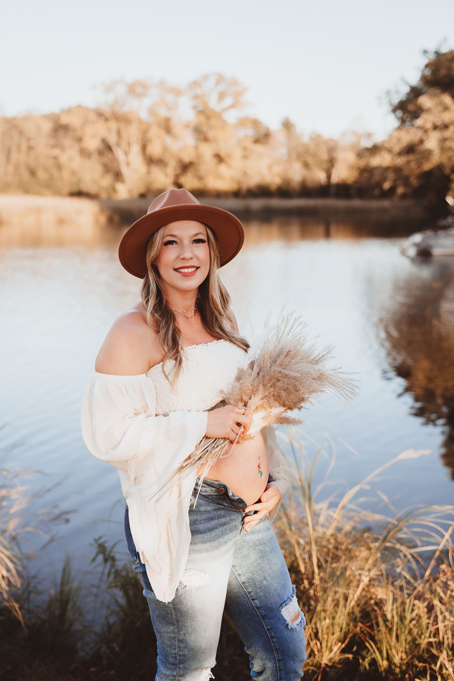 Radiant pregnant woman in a brown hat and white top holding dried pampas grass by a sunlit lake.