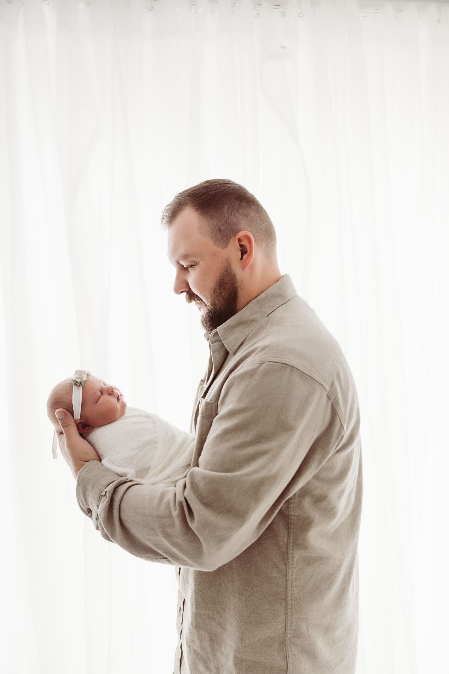 Smiling bearded father holding his swaddled newborn baby girl in a bright white studio.