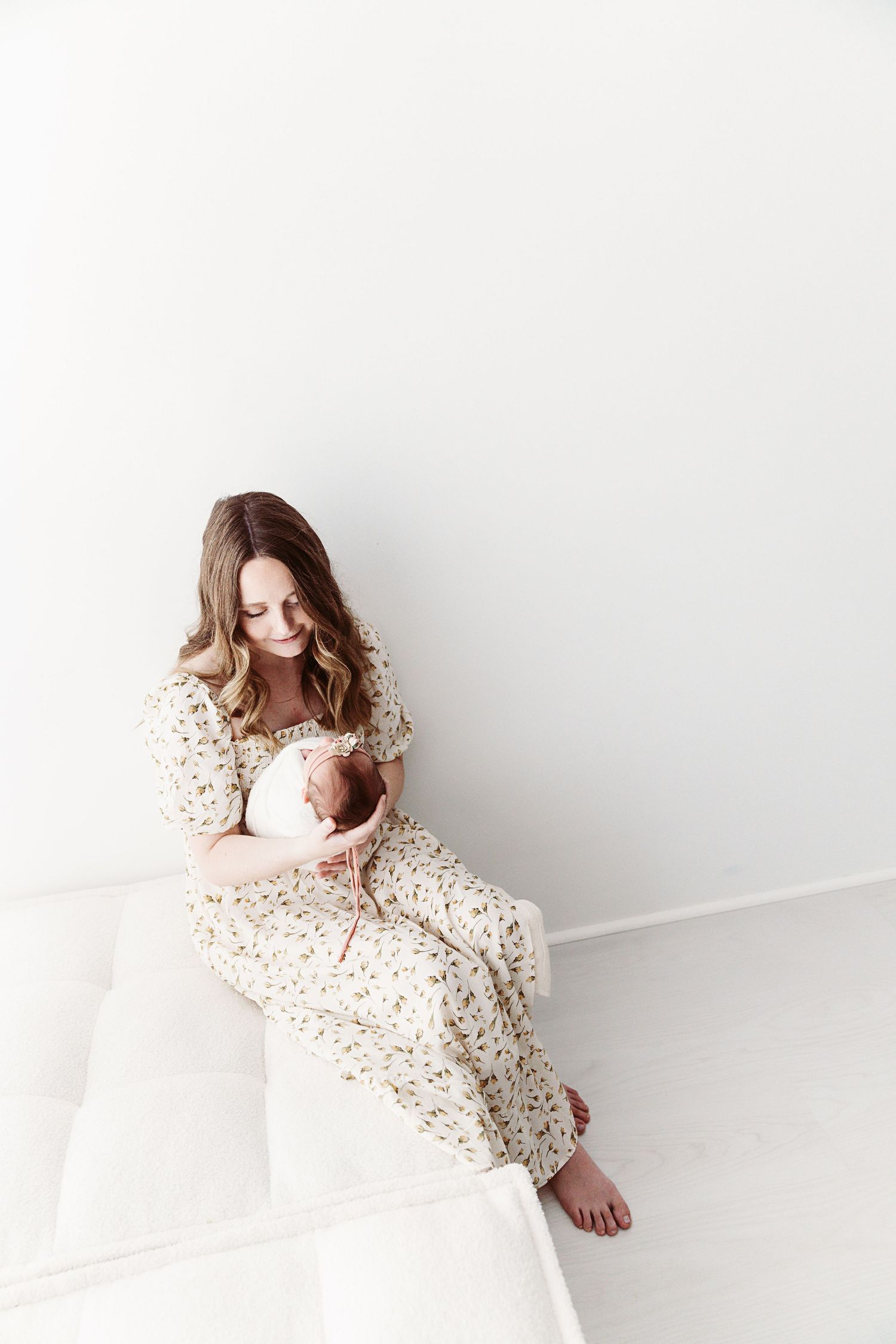 A mother in a floral dress sits on a white surface, gently holding her sleeping newborn baby.