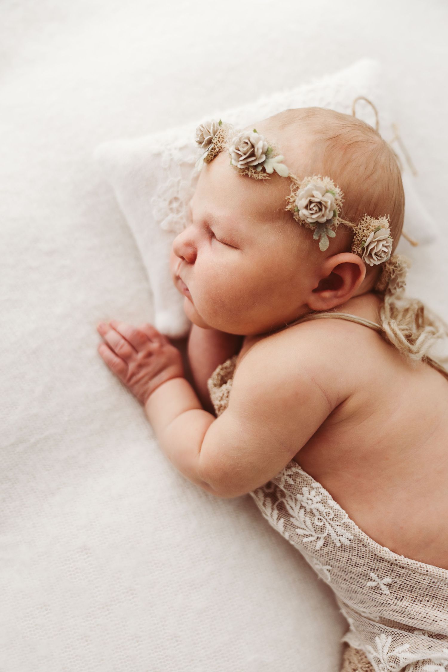 Peaceful newborn baby girl with floral headband sleeping on white blanket with lace wrap.
