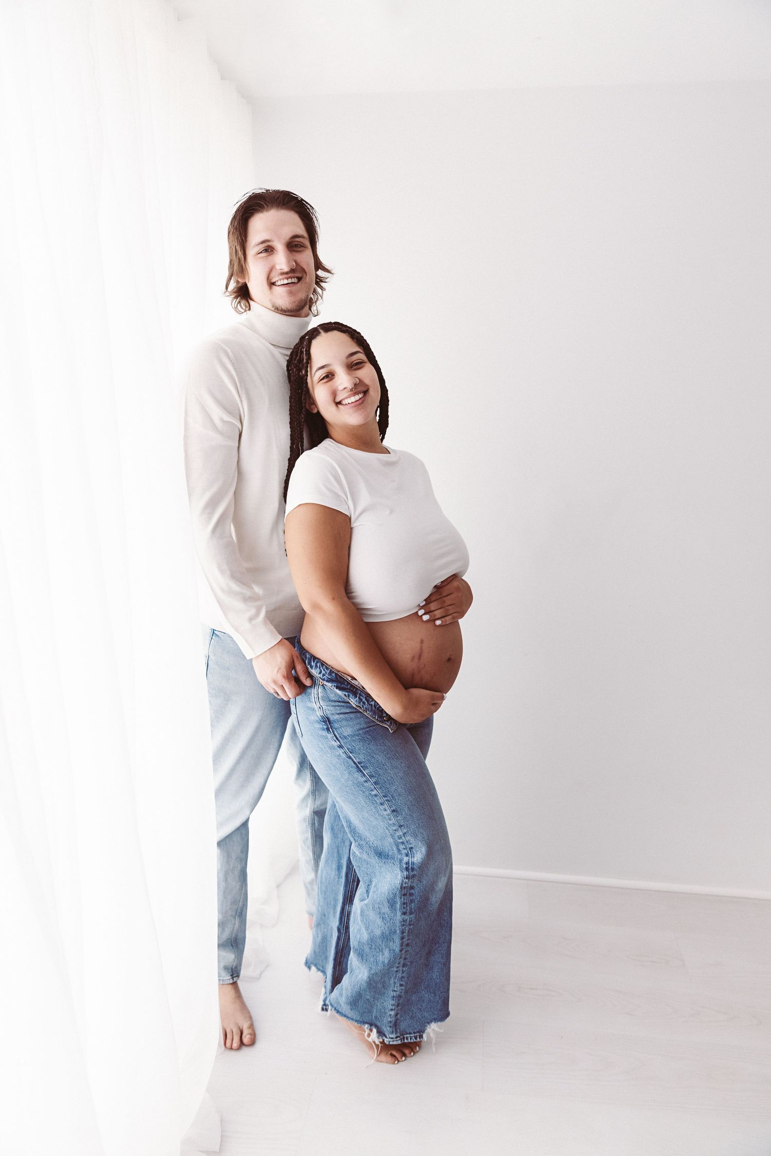 Happy couple expecting a baby, man embracing pregnant woman in white top and jeans in bright room.